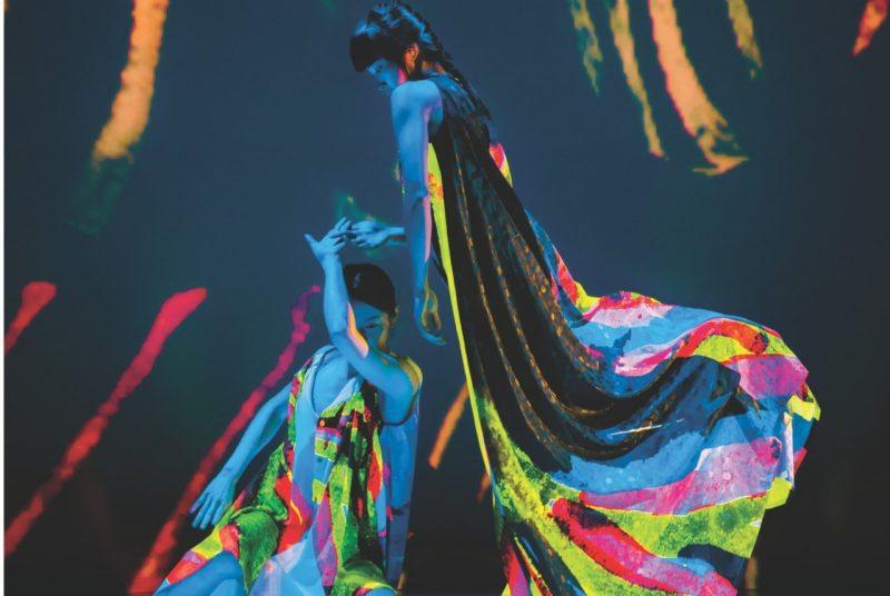 Two dancers with Cloud Gate Dance Theatre of Taiwan perform on stage wearing neon costumes that glows under a black light. One dancer sits while the other stands above her looking down. 